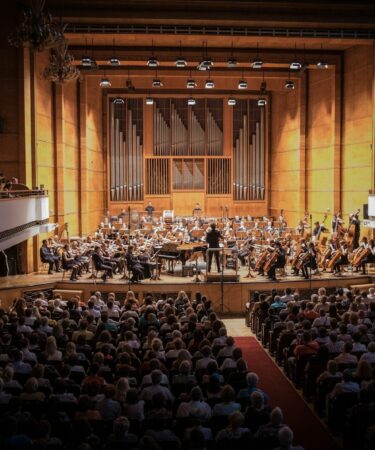 Public assis dans une salle de concert, face à un orchestre sur scène